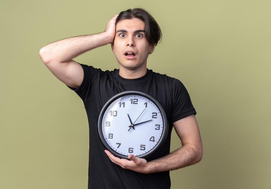 O Melhor Momento Para Validação é AGORA! 6 jovem bonito com medo de camiseta preta segurando um relogio de parede preso na cabeca isolada na parede verde oliva 141793 102851