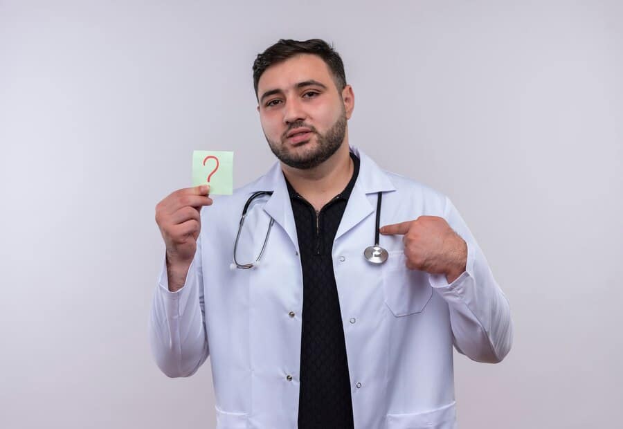 jovem medico barbudo vestindo jaleco branco com estetoscopio segurando um papel lembrete com ponto de interrogacao apontando com o dedo indicador para ele 141793 28164